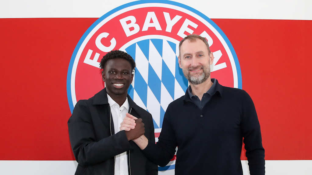 Bajung Darboe and Jochen Sauer shake hands in front of an FC Bayern logo.