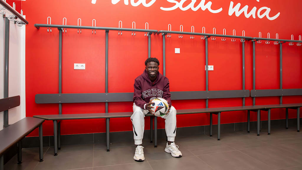 Bajung Darboe sits in the dressing room at the FC Bayern Campus stadium and holds a ball.