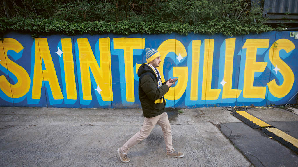A Union SG fan walks through the Saint-Gilloise neighbourhood in front of a wall covered in graffiti.