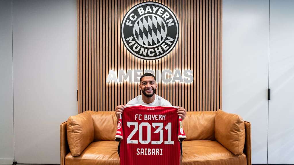 Ismael Saibari sitzt auf einem Sofa und hält lächelnd ein rotes Trikot mit der Aufschrift FC Bayern 2031 Saibari hoch