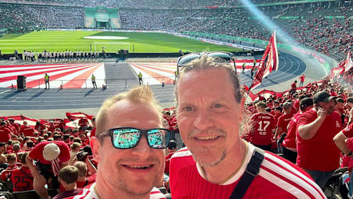 Zwei Männer in roten Bayerntrikots blicken in die Kamera, im Hintergrund kann man das Olympiastadion in Berlin erkennen.