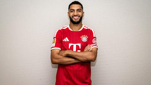 Ismael Saibari steht lächelnd mit verschränkten Armen im FC Bayern-Trikot an einer Wand.