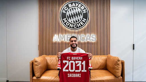 Ismael Saibari sitzt auf einem Sofa und hält lächelnd ein rotes Trikot mit der Aufschrift FC Bayern 2031 Saibari hoch