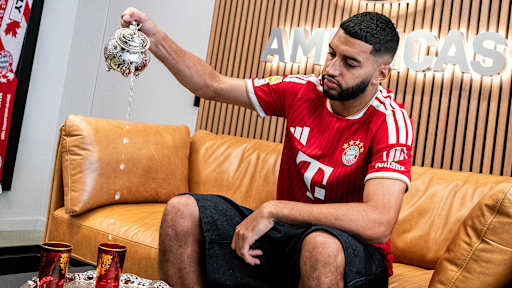 Ismale Saibari im roten FC Bayern-Trikot schenkt Tee aus einer silbernen Kanne in ein Glas