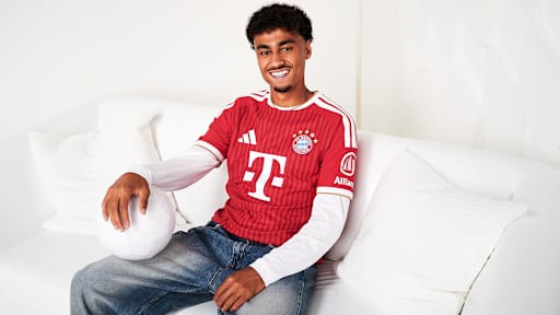 Nathaniel Brown sitzt im FC Bayern-Trikot auf einer weißen Couch und hat einen Ball im Arm