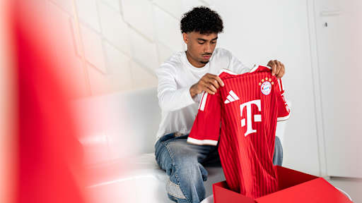 Nathaniel Brown sitzt auf einer weißen Couch und schaut sich ein FC Bayern Trikot an