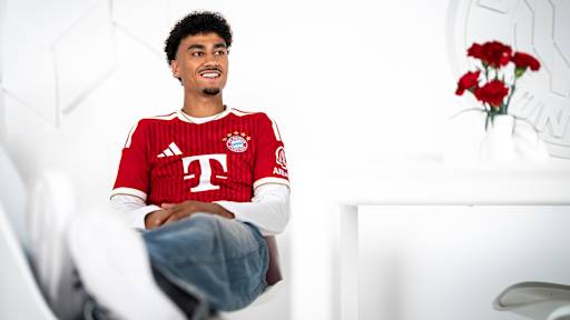 Nathaniel Brown sitzt im FC Bayern-Trikot auf einer weißen Couch