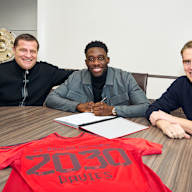 Max Eberl, Alphonso Davies und Christoph Freund sitzen an einem Tisch, Davies unterschreibt einen Vertrag