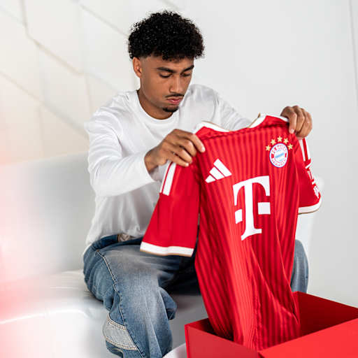 Nathaniel Brown sitzt auf einer weißen Couch und schaut sich ein FC Bayern Trikot an