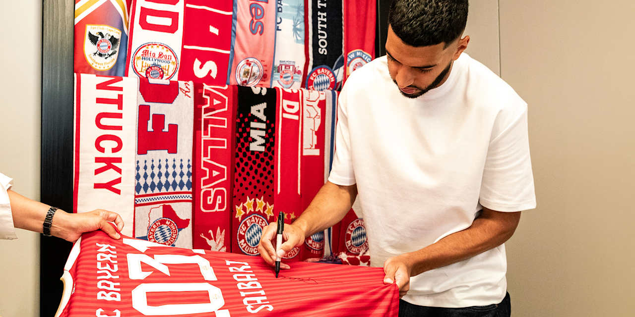 Ismael Saibari unterschreibt ein FC Bayern-Trikot mit der Zahl 2013 darauf vor aufgehängten Fan-Schals mi