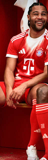 Harry Kane, Serge Gnabry und Konrad Laimer sitzen lachend in der Kabine und tragen das neue Heimtrikot des FC Bayern in Rot und Weiß mit stilisiertem M.