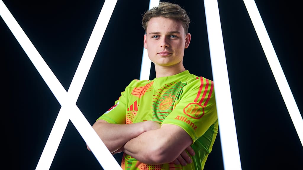 Jonas Urbig steht zwischen Leuchtröhren im FC Bayern-Trikot mit verschränkten Armen und schaut in die Kamera