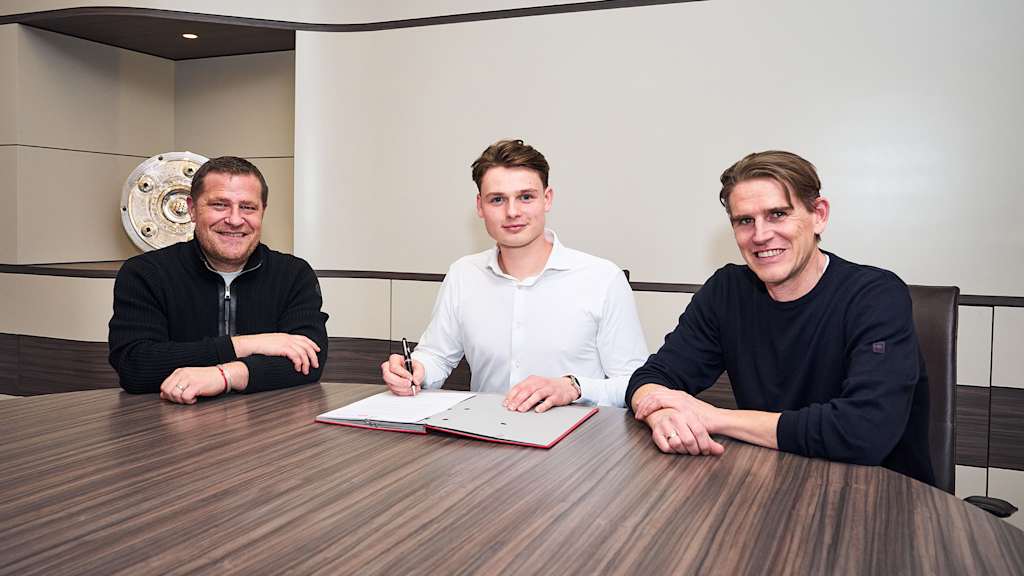 Max Eberl, Jonas Urbig und Christoph Freund sitzen an einem Tisch, Urbig unterschreibt seinen Vertrag beim FC Bayern