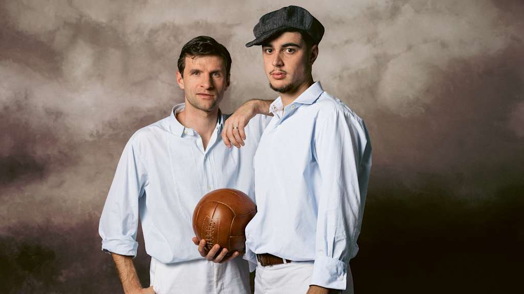 Thomas Müller und Aleks Pavlovic stehen nebeneinander in alter Spielkleidung des FC Bayern, Pavlovic hält einen Lederball in der Hand