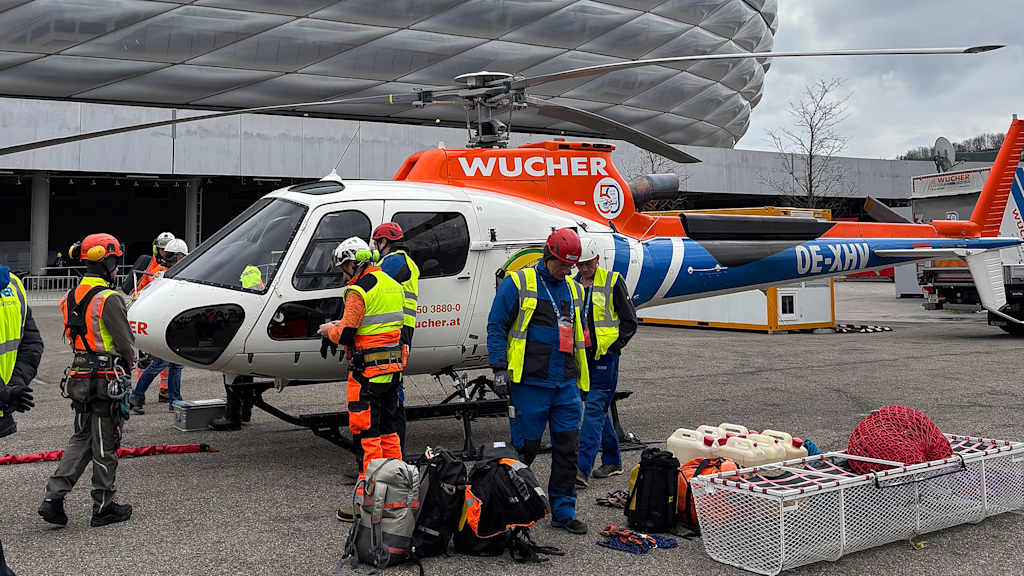 Arbeiter stehen vor einem Hubschrauber, im Hintergrund die Allianz Arena
