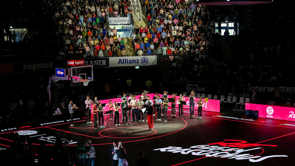 A youth orchestra plays on the basketball court, while the youth choir sings in the stands behind them.