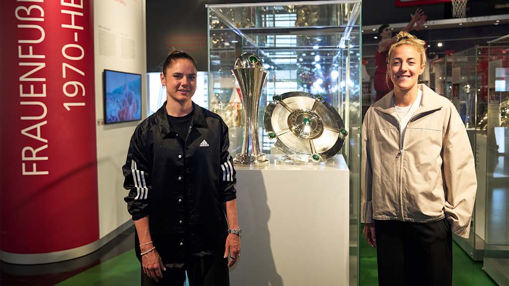 Sarah Zadrazil und Caro Simon neben der Vitrine im FC Bayern Museum mit den Double-Trophäen der FC Bayern Frauen aus der Saison 2024/25.