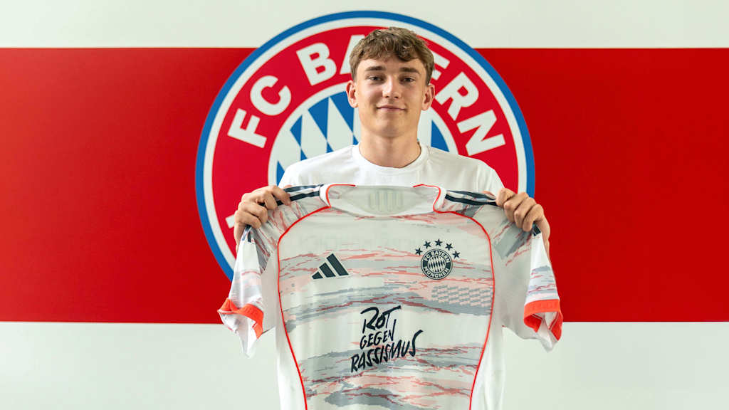 Artur Degraf posiert mit dem Trikot in der Hand vor dem FC Bayern Wappen.