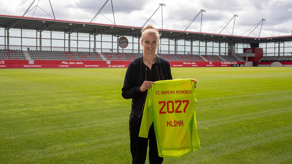 Anna Klink stands on the pitch of the stadium at the FC Bayern Campus and holds her goalkeeper jersey with the contract term 2027 to the camera.