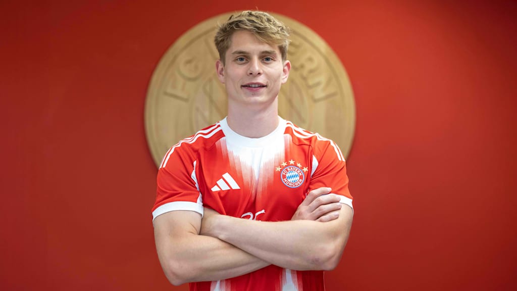 Anton Heinz posiert mit dem Trikot vor dem FC Bayern Wappen.
