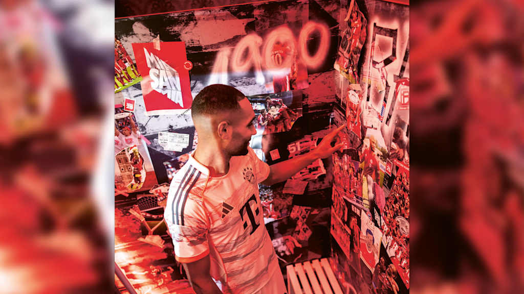 Jonathan Tah, in a white FC Bayern jersey, points to pictures on a wall