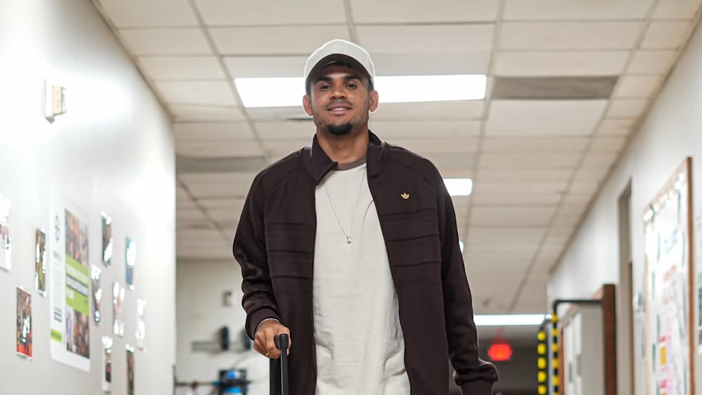 Luis Díaz reist in den USA an, hat eine braune Jacke an und lacht in die Kamera