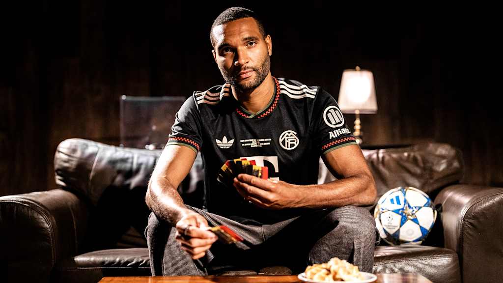 Jonahthan Tah sitzt im Champions League-Trikot des FC Bayern auf dem Sofa und hält Spielkarten in der Hand