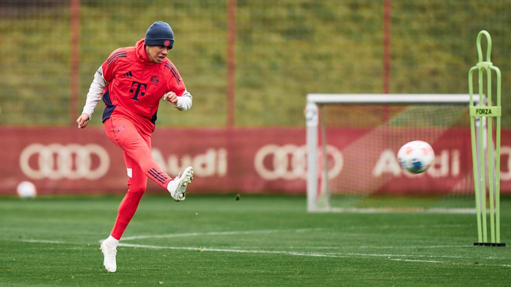 Jamal Musiala in roter Teamline und mit einer grauen Mütze schießt den Ball auf dem grünen Rasen.