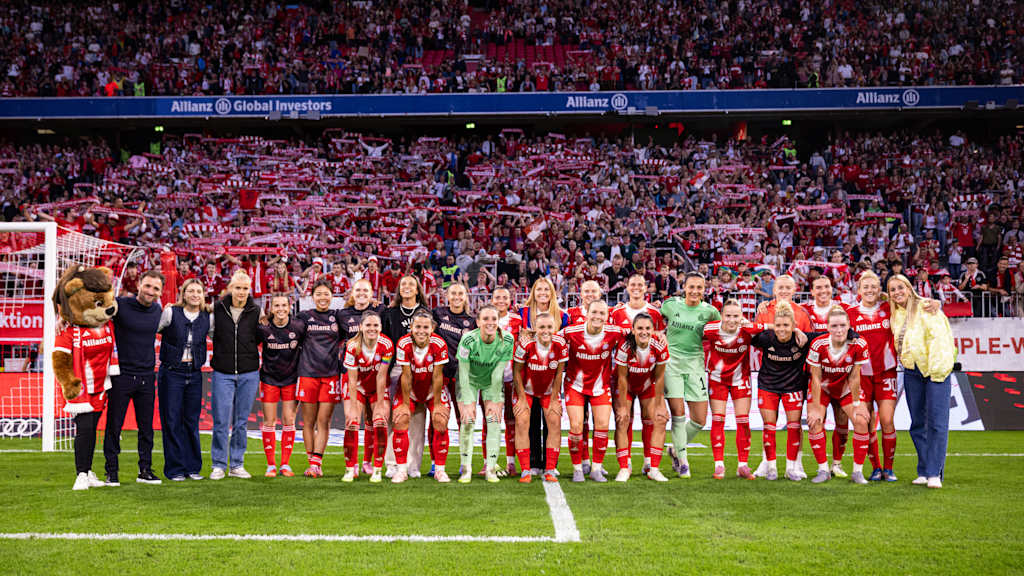 The FC Bayern Women in a group photo in front of the south stand of the Allianz Arena with fans in the background.
