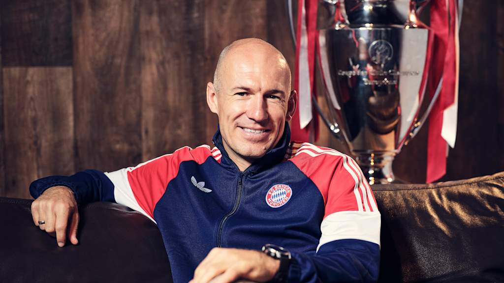 Arjen Robben grinst in einer Trainingsjacke des FC Bayern auf einer Couch.