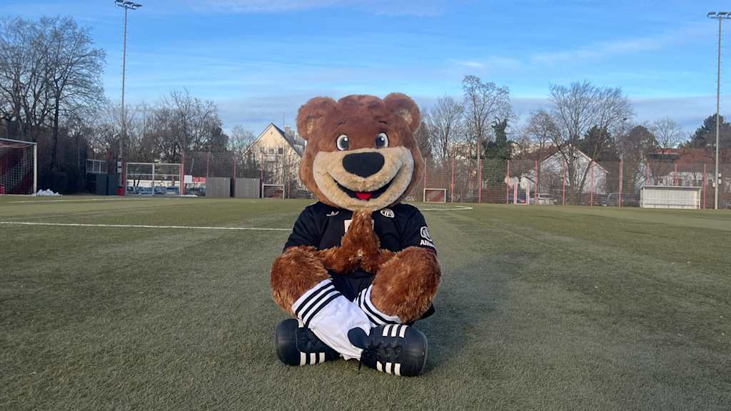 Berni auf dem Kunstrasenplatz in einem Schneidersitz und gefalteten Händen.