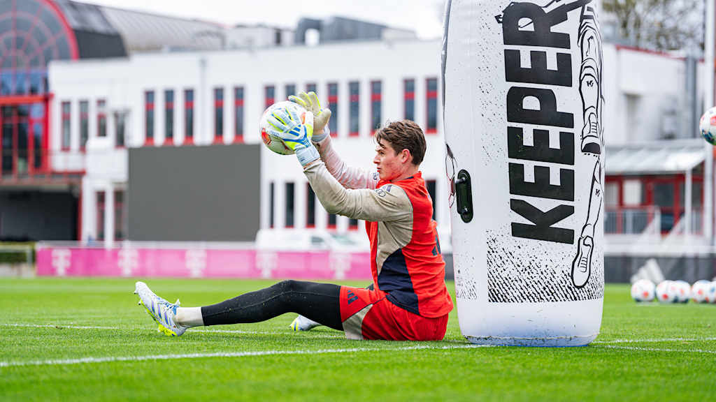 Jonas Urbig hält im Training des FC Bayern einen Ball im Sitzen in den Händen