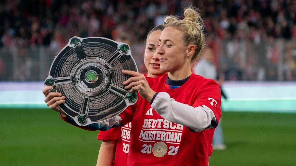 Carolin Simon mit einer Papierversion der Meisterschale und einem T-Shirt mit der Aufschrift 'Deutsche Meisterinnen 2026'. Hinter ihr steht Giulia Gwinn.