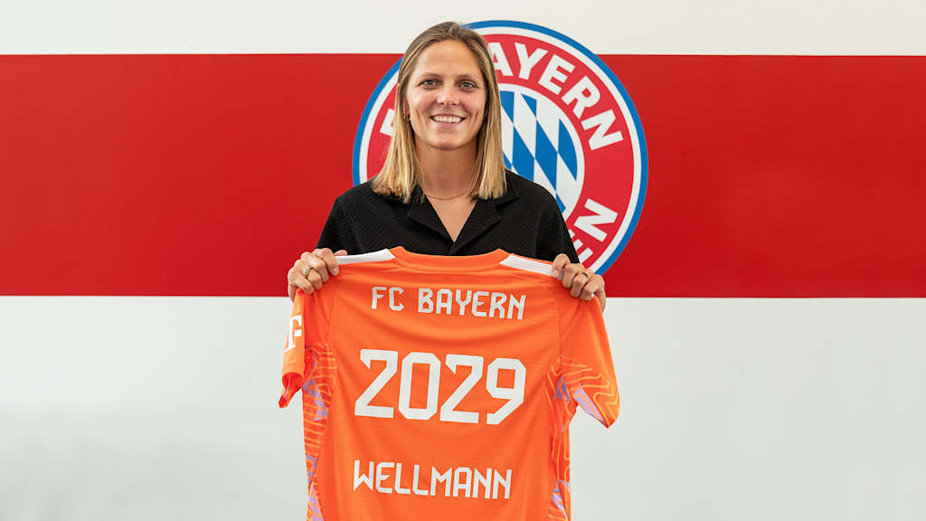 Anna Wellmann steht vor einer Wand mit Bayern Logo und hält ihr Trikot.