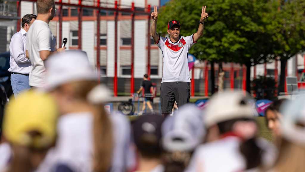 Olympiasieger Michael Greis hält vor Teilnehmern des Grundschulwettbewerbs an der Säbener Straße an beiden Händen jeweils zwei Finger hoch