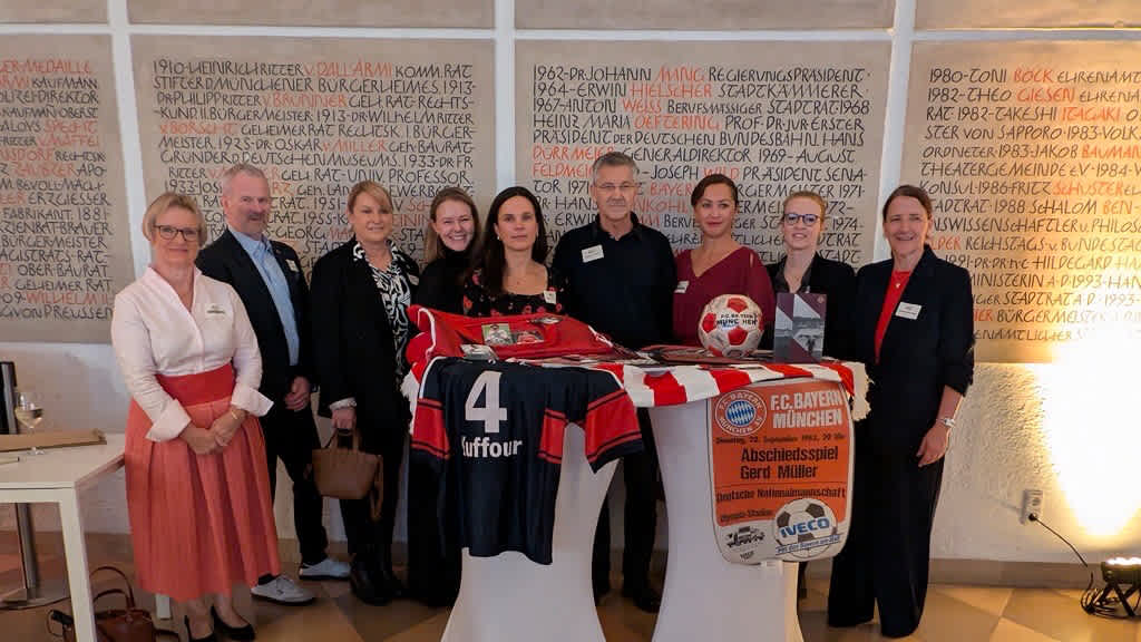 Herbert Hainer steht mit Verantwortlichen zum Jubiläum der Alzheimergesellschaft an einem Stehtisch und zeigt den Erinnerungskoffer des FC Bayern