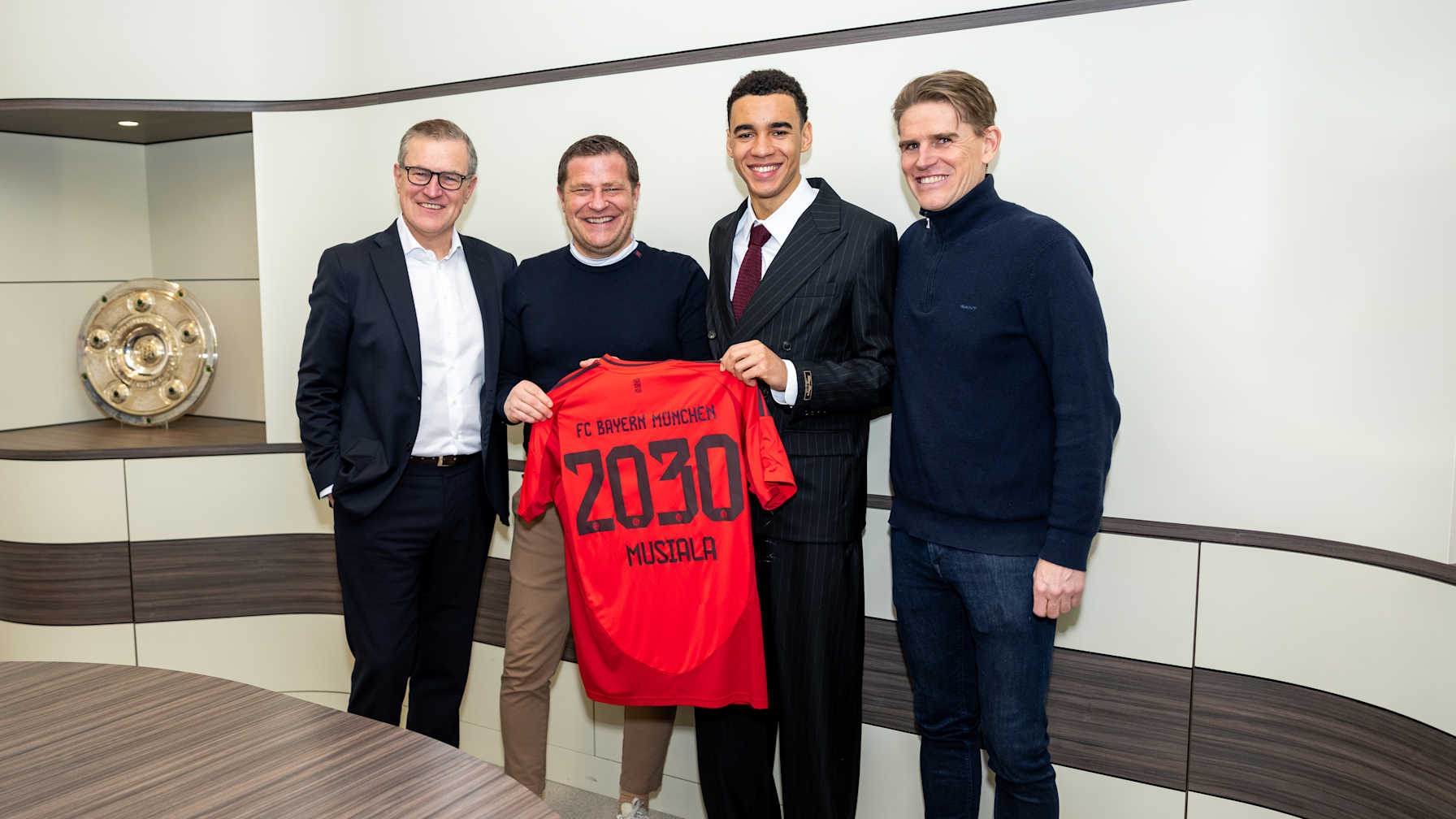 Jan-Christian Dreesen, Max Eberl, Jamal Musiala und Christoph Freund stehen nebeneinader und halten ein Bayern-Trikot hoch mit 2030 Musiala