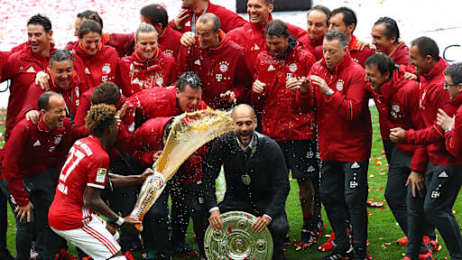 Pep Guardiola wird mit Bier überschüttet als er mit der Meisterschale und dem Trainerteam posiert.