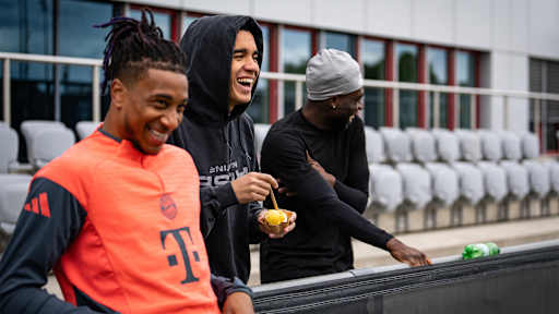 Michael Olise, Jamal Musiala und Alphonso Davies beobachten lachend das FC Bayern-Training vor Bundesliga-Spiel bei TSG Hoffenheim.