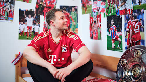 Manuel Neuer sitzt in einem FC Bayern-Trikot in einem Kinderbett mit Bayern-Bettwäsche in einer Nachbildung eines Thomas Müller-Kinderzimmers.