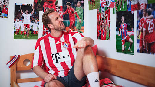 Harry Kane sitzt in einem FC Bayern-Trikot in einem Kinderbett mit Bayern-Bettwäsche in einer Nachbildung eines Thomas Müller-Kinderzimmers.