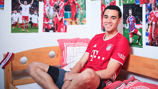 Jamal Musiala sitzt in einem FC Bayern-Trikot in einem Kinderbett mit Bayern-Bettwäsche in einer Nachbildung eines Thomas Müller-Kinderzimmers.