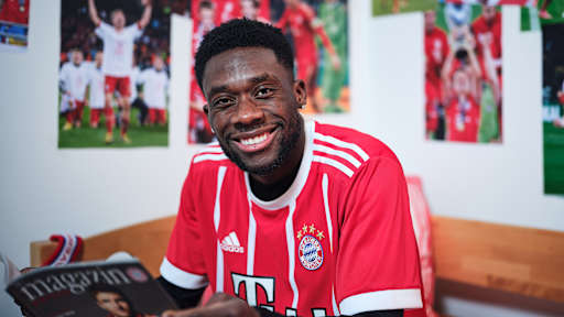 Alphonso Davies sitzt in einem FC Bayern-Trikot in einem Kinderbett mit Bayern-Bettwäsche in einer Nachbildung eines Thomas Müller-Kinderzimmers.