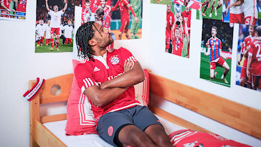 Serge Gnabry sitzt in einem FC Bayern-Trikot in einem Kinderbett mit Bayern-Bettwäsche in einer Nachbildung eines Thomas Müller-Kinderzimmers.