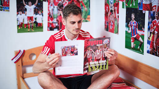 Josip Stanisic sitzt in einem FC Bayern-Trikot in einem Kinderbett mit Bayern-Bettwäsche in einer Nachbildung eines Thomas Müller-Kinderzimmers.
