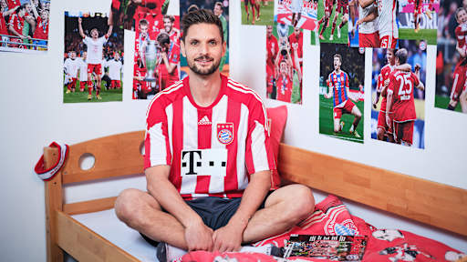 Sven Ulreich sitzt in einem FC Bayern-Trikot in einem Kinderbett mit Bayern-Bettwäsche in einer Nachbildung eines Thomas Müller-Kinderzimmers.