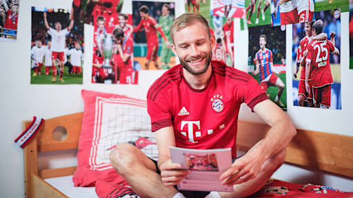 Konrad Laimer sitzt in einem FC Bayern-Trikot in einem Kinderbett mit Bayern-Bettwäsche in einer Nachbildung eines Thomas Müller-Kinderzimmers.
