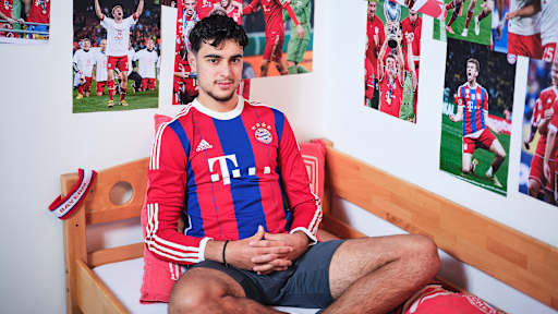 Aleksandar Pavlovic sitzt in einem FC Bayern-Trikot in einem Kinderbett mit Bayern-Bettwäsche in einer Nachbildung eines Thomas Müller-Kinderzimmers.