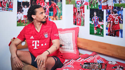 Leroy Sane sitzt in einem FC Bayern-Trikot in einem Kinderbett mit Bayern-Bettwäsche in einer Nachbildung eines Thomas Müller-Kinderzimmers.