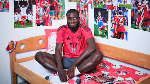 Dayot Upamecano sitzt in einem FC Bayern-Trikot in einem Kinderbett mit Bayern-Bettwäsche in einer Nachbildung eines Thomas Müller-Kinderzimmers.